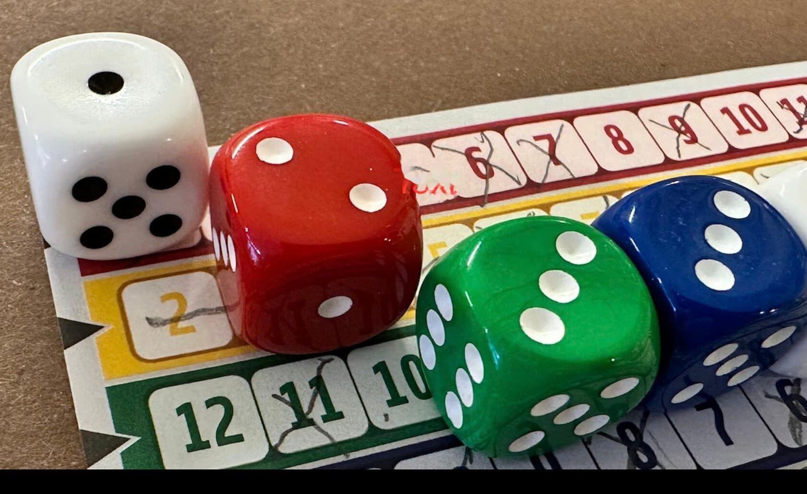 Vier bunte Würfel (weiß, rot, grün, blau) auf einem Spielplan mit durchgestrichenen Zahlenfeldern
